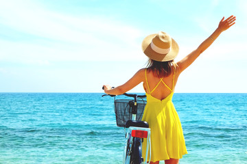 Fototapeta na wymiar Young carefree woman in bright yellow dress with bicycle at ocean beach. Unrecognizable female wearing broad brim straw hat biking on sandy sea shore on sunny day. Copy space, background.