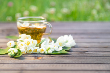 Fototapeta na wymiar Glass cup with hot fragrant jasmine tea on a wooden background. Around a cup leaflets and flowers of a jasmine are spread out. On a background a green lawn with the blossoming clover
