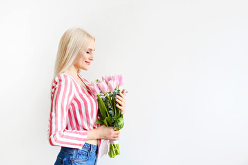 Fototapeta na wymiar Studio portrait of gorgeous young blonde woman with long straight hair wearing pink stripe crop top shirt, holding bouquet of many tender pink tulips. White isolated background, copy space, close up.