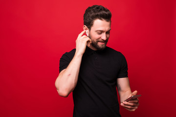 Portrait of happy bearded man singing and listening to music with cell phone and wireless earphones isolated over red wall