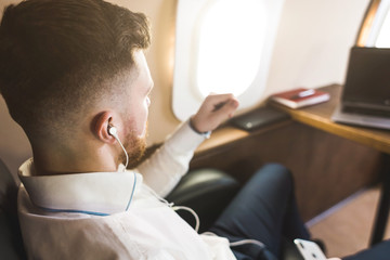 Fototapeta premium Young attractive and successful businessman is resting and listening to music works while sitting in the chair of his private business plane