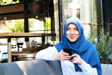 Young beautiful caucasian woman wearing traditional muslim headscarf in hipster coffee shop with big full length windows. Female in blue hijab at cozy cafe. Background, copy space, close up portrait.