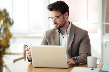 Handsome young business man using his smartphone while working with laptop in the office.