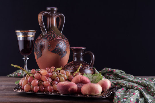 Still life with a wine glass next to ceramic vessels and grapes with peaches on a beautiful tin tray on a wooden table on a black background