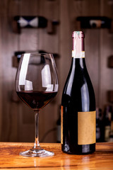 Glass and bottle with delicious red wine on wooden table. In the background Red wine bottles stacked on wooden racks shot with limited depth of field.