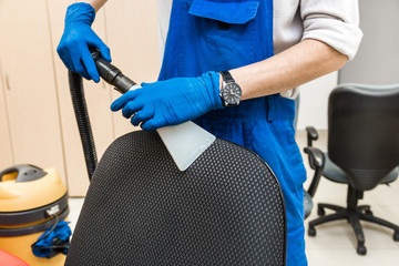 Young man in workwear and rubber gloves cleans the office chair with professional equipment.