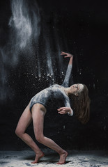 Obraz na płótnie Canvas A young beautiful girl in a swimsuit and a translucent skirt emotionally dancing on a black background in clouds of white dust