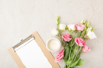 Obraz na płótnie Canvas Top view with blank paper clipboard, rose and white blooming flowers, candle and office accessories. Flat lay with copy space. Home office desk in rose and white colors