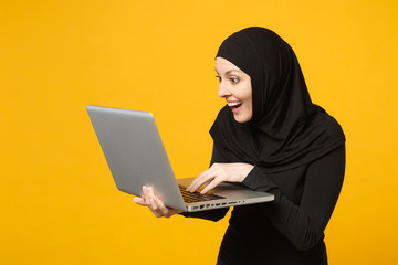 Young arabian muslim employee woman in hijab black clothes hold and work laptop pc computer isolated on yellow wall background, studio portrait. People religious lifestyle concept. Mock up copy space.