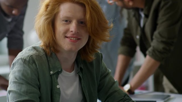 Portrait shot of young male designer with thick red hair sitting at table writing project notes, posing and confidently smiling for camera, with multiracial group of colleagues talking in background