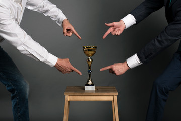 Two businessmans is showing by index fingers on a golden cup award trophy on a chair. Struggle for...