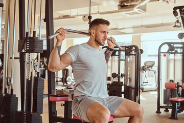 Young tattoed man is doing exercise for back on training apparatus in gym.