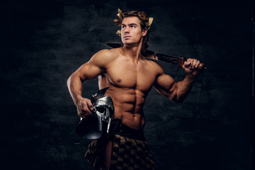 Fototapeta premium Attractive powerful man in wreath is posing on grey background and holds helmet and sword in his hands.