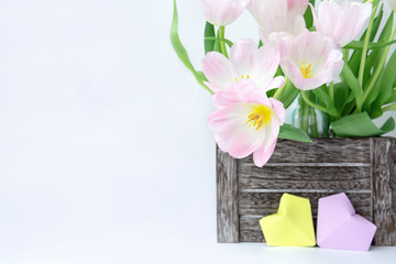 A bouquet of pink tulips in a wooden box and two paper hearts of yellow and lilac color on a white background.