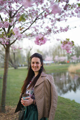 Young woman drinking coffee from a paper cup wearing emerald color skirt - Colorful sakura cherry blossom in a park in Riga, Eastern European capital city of Latvia