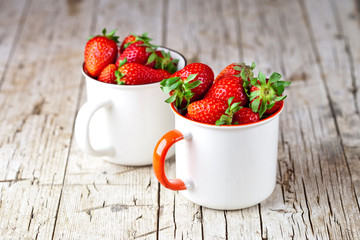 Organic red strawberries in two white ceramic cups on rustic wooden background.