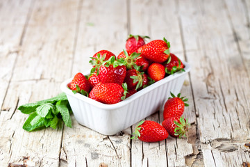 Fresh red strawberries in white bowl and mint leaves on rustic wooden background.