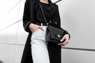 Young stylish woman in a fashionable long black coat in vintage white jeans in a black trendy t-shirt with a leather stylish black handbag is standing outdoors. Modern women's fashion. Spring-Autumn.
