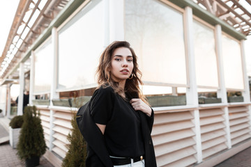 Urban beautiful young woman in a black coat in a fashionable t-shirt in white jeans with a pose posing near a vintage wooden building outdoors. European girl model enjoys spring day. Youth style.