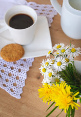Obraz na płótnie Canvas View of a white porcelain cup of black coffee, bunch of dandelions and daisies, a cookie on a wooden table with a vintage doily illuminated by sun light. Good morning coffee concept