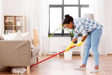 housework and housekeeping concept - african american woman or housewife with mop cleaning floor under sofa at home