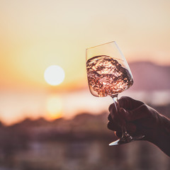 Glass of rose wine in mans hand with sea and sunset at background, close-up, square crop. Summer...