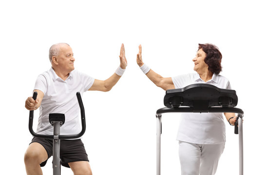Senior man on a stationary bike and a senior woman on a treadmill high-fiving each other