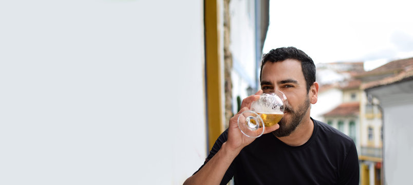 Isolated Hispanic latin arab indian bearded handsome guy drinking beer with a a blurred colonial city background, and plenty of horizontal copy space. Ads, banner, blank space and advertising concept