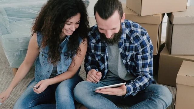 Young couple having rest after moving stuff to their new apartment, sitting on floor, watching comedy movie.