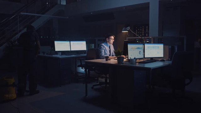 Working Late at Night in the Office: Businessman Uses Desktop Computer, Analyzing, Using Documents, Solving Problems, Finishing Important Project. Diligent Ambitious Worker. Cleaning Man Vacuums Floor