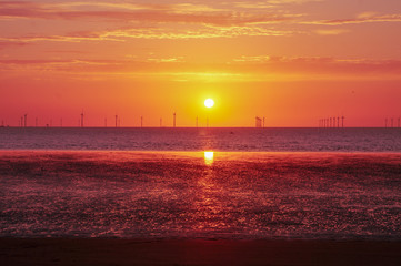 Fototapeta premium Wind turbines and beach at sunset. Reflecting low tide from the vivid sky above. Strong orange, reds and purples. Cloudy sky and the ocean