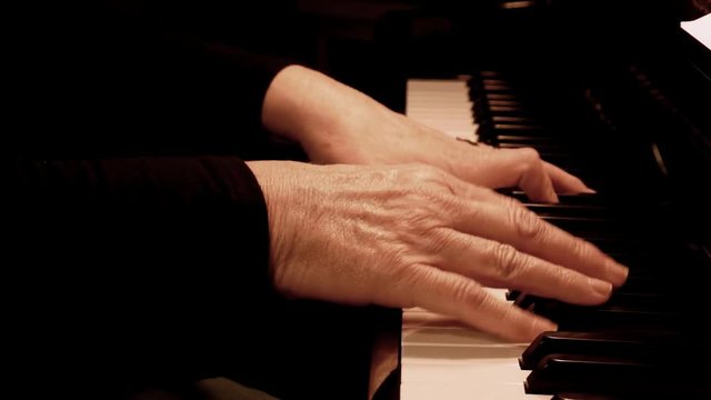 Senior woman's hands playing piano. Close up side view of elderly hands and fingers playing a song on the keys of an upright shiny black piano.
