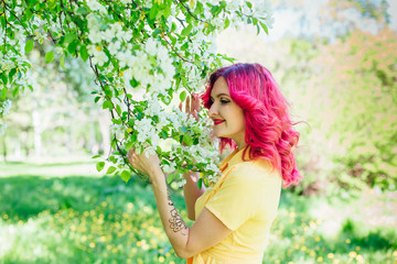 Beautiful and young woman with bright red hair and ref lips standing next to a blooming apple tree in a yellow dress.
