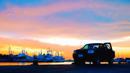 Fototapeta na wymiar Silhouette pick up car parking on viewpoint with blurred background of many cargo ships docked at port on riverside against colorful sunrise sky in evening time