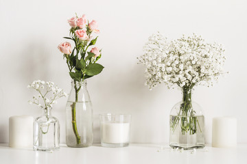 Composition with white and pink flowers in glass bottles and candles on table wall background