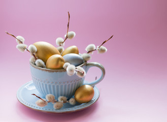Easter eggs of gold and silver color with willow branches in a porcelain mug on a pink background. Front view. Copy space