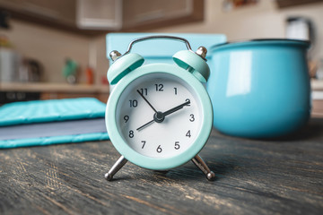 Blue metal alarm clock on the table with a mug and a diary on the background of the kitchen. Time...