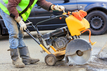 A worker in a green reflective vest cuts a piece of bad asphalt with a gasoline cutter on the carriageway.