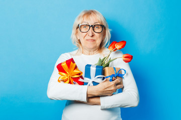 Old woman with a happy facial expression is holding presents and a bouquet of tulips in her hands against a blue background. Mothers Day, congratulation, birthday and holiday concept.