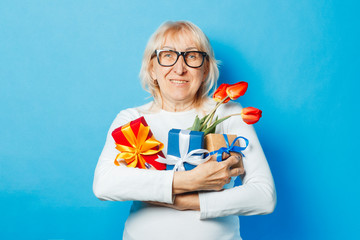 Old woman with a happy facial expression is holding presents and a bouquet of tulips in her hands against a blue background. Mothers Day, congratulation, birthday and holiday concept.