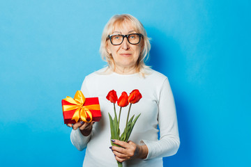 Old woman with a happy facial expression is holding presents and a bouquet of tulips in her hands against a blue background. Mothers Day, congratulation, birthday and holiday concept.