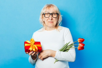 Old woman with a happy facial expression is holding presents and a bouquet of tulips in her hands against a blue background. Mothers Day, congratulation, birthday and holiday concept.