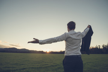 Businessman standing in green meadow looking towards the sunset
