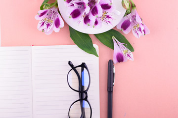 Fragrant tea in a white cup, beautiful flowers, a pink background, a notebook with a pen and glasses.