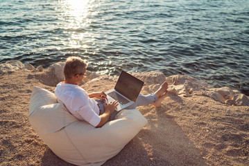 Senior man working on his laptop lying on deck chair on the beach during sunset