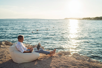 Senior man working on his laptop lying on deck chair on the beach during sunset