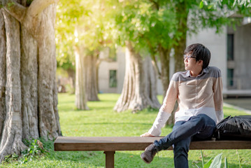 Naklejka na ściany i meble Young Asian man university student with glasses sitting on wooden bench in the college garden. Campus lifestyle in high school. Education opportunity or scholarship concepts