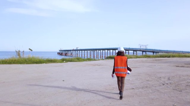 Giovane ingegnere donna con gilet arancione e caschetto bianco va a lavorare al pontile sulla spiaggia di fronte all'oceano.
