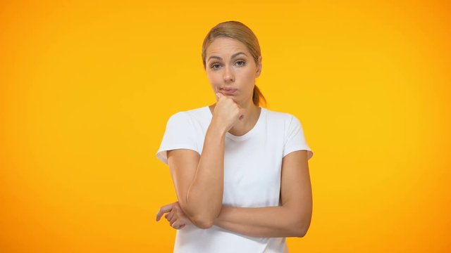 Thoughtful female with hand on chin standing on orange background, hesitation