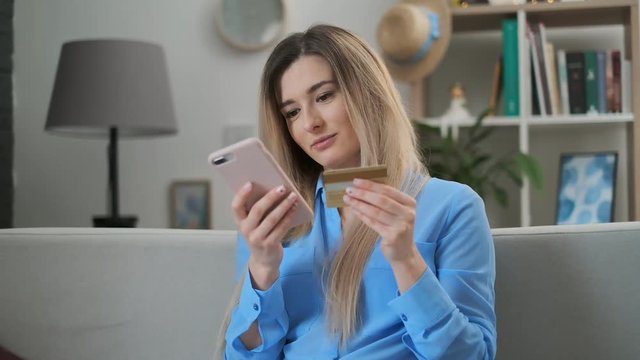 Girl buys gifts online at home with card and phone app. Woman sits on sofa, holding smartphone entering card number. Easy e-commerce transaction, booking, shooping online.